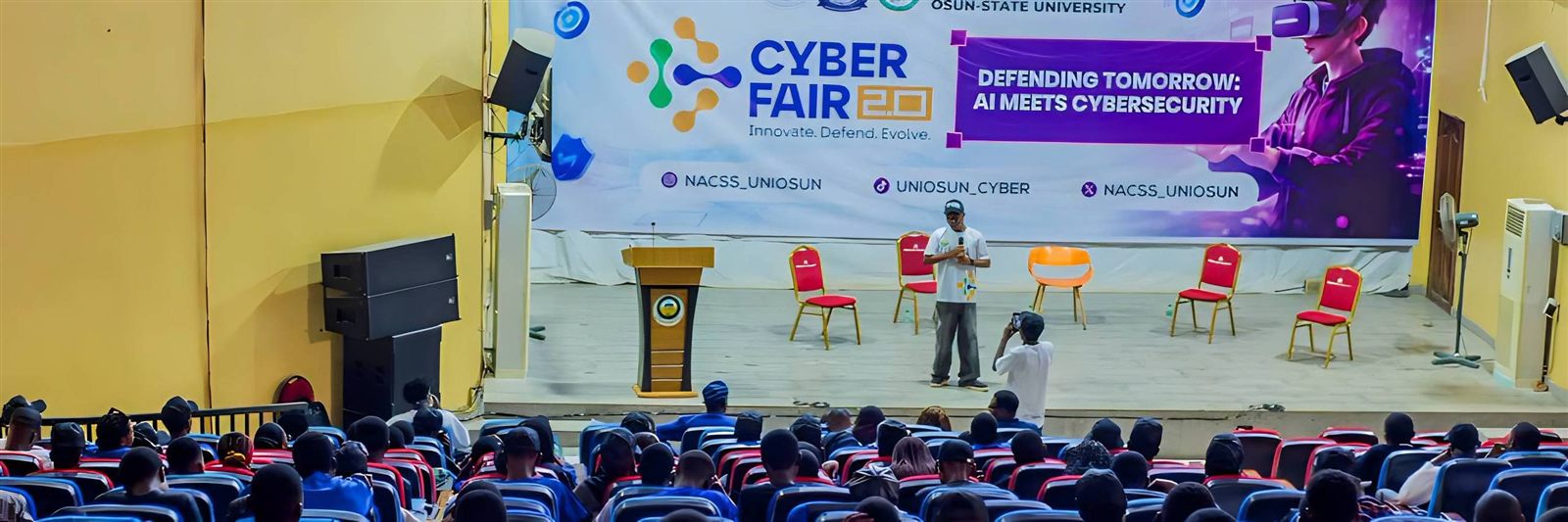 A speaker on stage at Cyber Fair 2.0 in front of a full auditorium, under a banner reading Defending Tomorrow: AI Meets Cybersecurity
